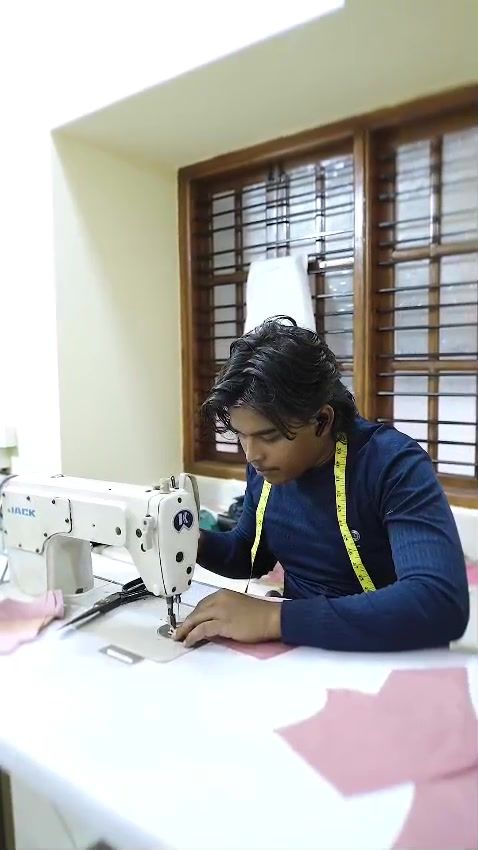 A tailor at an industrial sewing machine in the SLB studio, measuring tape around his neck